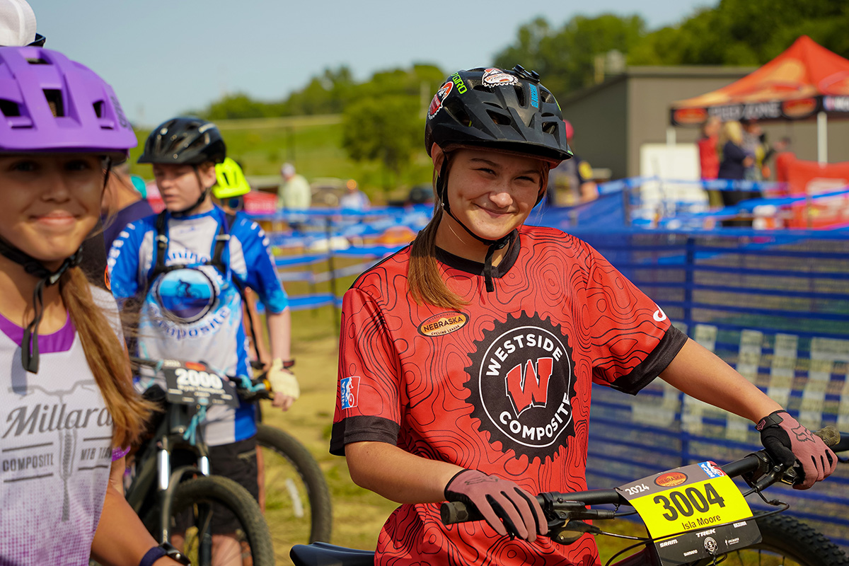 A girl in a red Westside Composite cycling jersey and helmet smiles while holding her bike, surrounded by other young cyclists at a biking event. Blue fencing and tents are visible in the background.