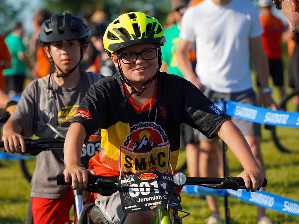 Jeremiah on his bike wearing a helmet and numbered jersey smiles as he prepares for a race. Other participants and blue event tape are visible in the background.