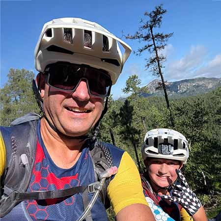Two people wearing biking helmets, sunglasses, and biking gear smile for a selfie outdoors. Behind them are green trees and a distant mountain under a blue sky.