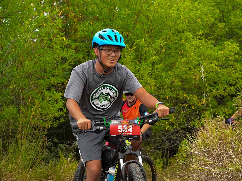 jaxson 1 A smiling Jaxson wearing a blue helmet and glasses rides a mountain bike with a race number 544 on the handlebar, on a trail surrounded by green foliage. Another rider is partially visible behind him.