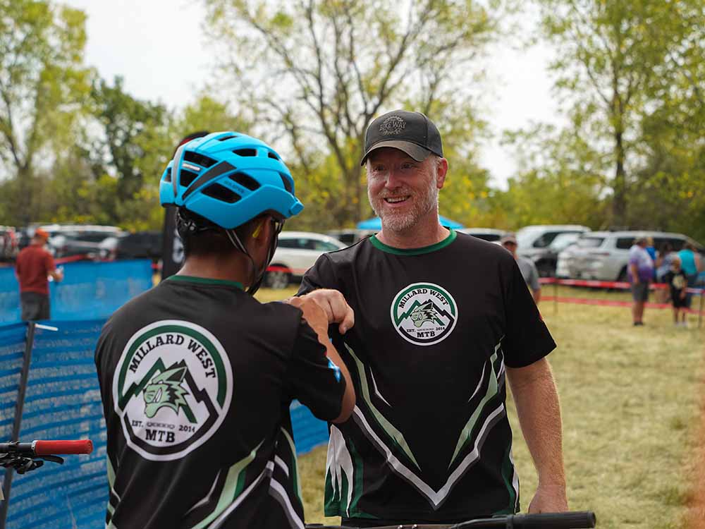 Two people wearing matching Millard West MTB jerseys greet each other with a fist bump at an outdoor biking event. Jaxson wears a blue helmet; cars and people are visible in the background.